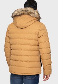 Doudoune beige avec une capuche bordée de fourrure, présentant des sections matelassées horizontales et des poches latérales. Matière texturée pour la chaleur.