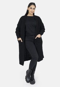 Schwarzer, langer Cardigan mit strukturiertem Strickdesign, offener Vorderseite und zwei vorderen Taschen, getragen über einem schwarzen Oberteil und einer schwarzen eng anliegenden Hose.