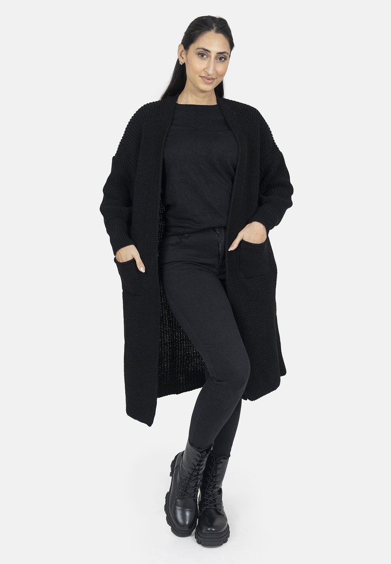 Schwarzer, langer Cardigan mit strukturiertem Strickdesign, offener Vorderseite und zwei vorderen Taschen, getragen über einem schwarzen Oberteil und einer schwarzen eng anliegenden Hose.