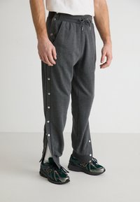 Graue Sporthose mit seitlichen Druckknöpfen, elastischem Bund und vorderen Taschen. Kombiniert mit grünen und schwarzen Sportschuhen.