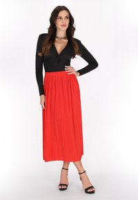 Schwarzes Langarmoberteil mit tiefem V-Ausschnitt und geknotetem Detail, kombiniert mit einem langen, plissierten roten Rock. Schwarze Riemchen-Heels runden den Look ab.