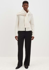 Frau trägt eine weiße, knöpfbare Strickjacke mit einem passenden Schal über den Schultern, schwarze weite Hosen und schwarze spitze Schuhe.