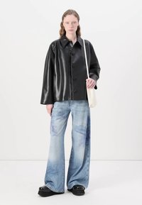 Chaqueta negra de cuero sintético con botones y un corte relajado, combinada con jeans de pierna ancha de color azul claro que presentan un sutil patrón de tie-dye y zapatos de plataforma negros.