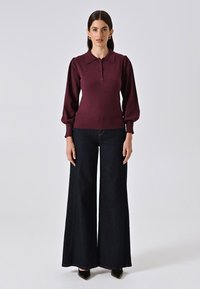 Top in magenta a maglia con colletto, tre bottoni e maniche a palloncino, abbinato a pantaloni larghi in denim scuro a vita alta e tacchi neri.