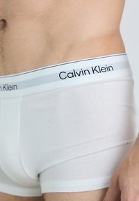 Bijeli pamučni bokserice s elastičnom trakom u struku koja ima sive naglaske i crni "Calvin Klein" logo. Glatka tekstura i prilagođeni dizajn.