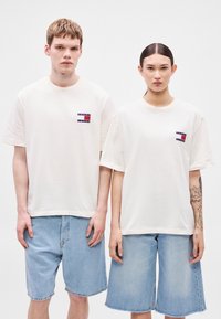 T-shirts brancas de algodão com mangas curtas, com um pequeno logótipo retangular no peito. Combinadas com calções de ganga azul claro.