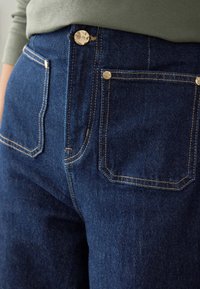 Jeans in denim blu scuro con vita alta, due tasche posteriori con cuciture decorative e accessori in metallo lucido.