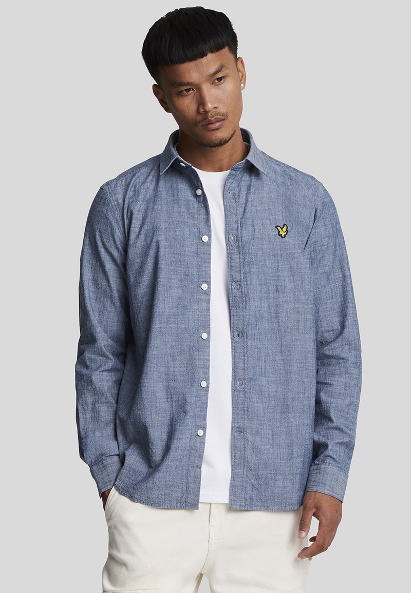 Btn-up skjorte i lyseblå chambray-stof med spidse kraver, lange ærmer og et gult broderet logo på brystet.