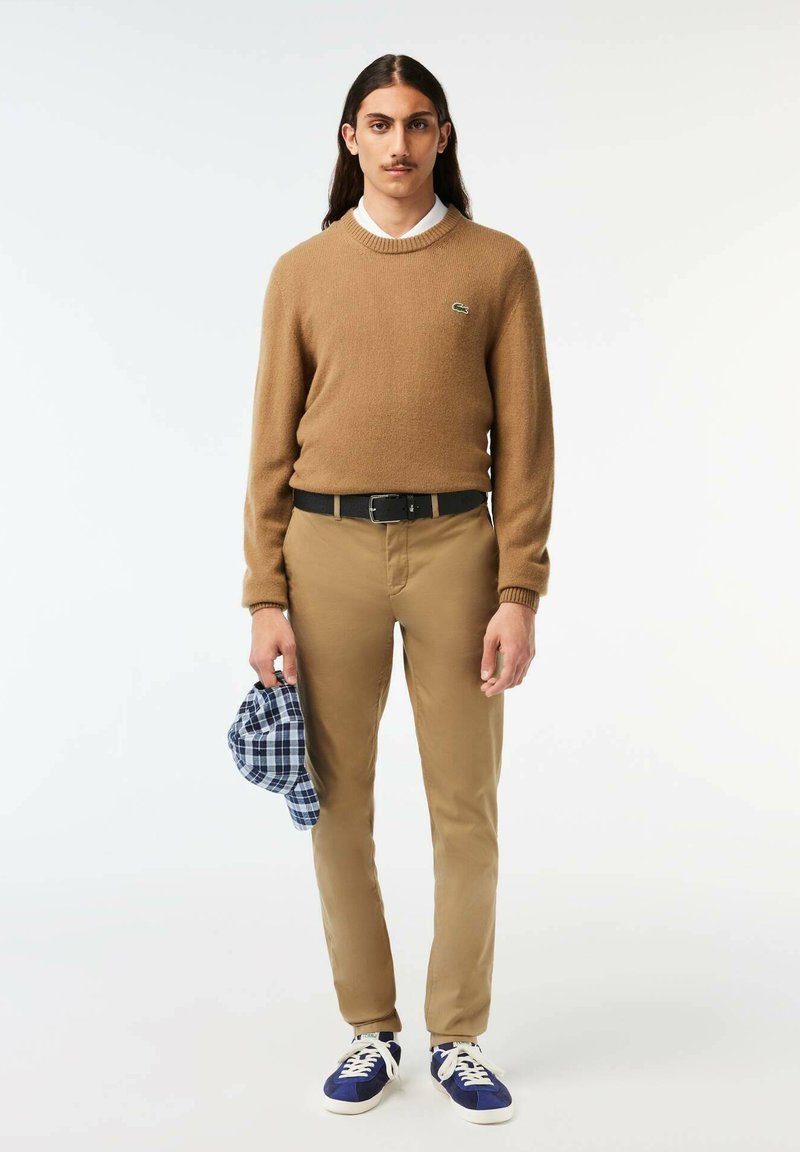 Brauner Strickpullover mit rundem Halsausschnitt, beigefarbene Chinos und blaue Sneakers. Hält eine blaue karierte Mütze. Einfaches, lässiges Design.