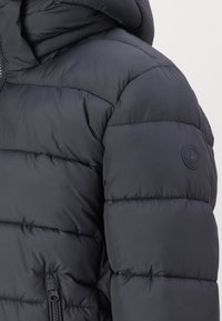 Jachetă puf din material închis la culoare, cu glugă, design matlasat și o eticheta rotundă cu logo pe umărul stâng. Buzunar cu fermoar pe lateral.