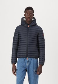 Dark navy puffer jacket with horizontal quilted design, front zipper, and adjustable hood. Features orange zipper pull and logo accent.