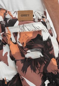 Pamut nadrág barna, fehér és narancssárga ló mintával, a hátsó derékrészén egy bézs műbőr címkével, rajta az "OBEY" felirattal.