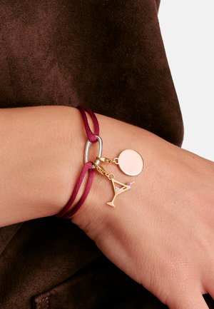 Handgelenk mit einem dunkelroten Kordelarmband, das einen silbernen Verschluss und zwei goldene Anhänger trägt: eine runde Scheibe und ein Martini-Glas mit einem pinkfarbenen Edelstein-Oliven.
