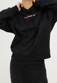 Czarna bluza z kapturem z przednią kieszenią typu kangur, sznurkami regulacyjnymi i napisem „LazyDreamer” w różowym kolorze. Miękka tkanina z prążkowanymi mankietami i obszyciem na dole.