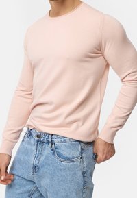 Svetle růžový svetr s dlouhým rukávem a kulatým výstřihem, vyrobený z měkké látky, v kombinaci s modrými denimovými džínami s klasickým střihem a vybledlým efektem.