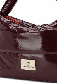 Bolso brillante color burdeos con forma redondeada, presenta una cremallera naranja y una etiqueta blanca con texto negro, hecho de material brillante.