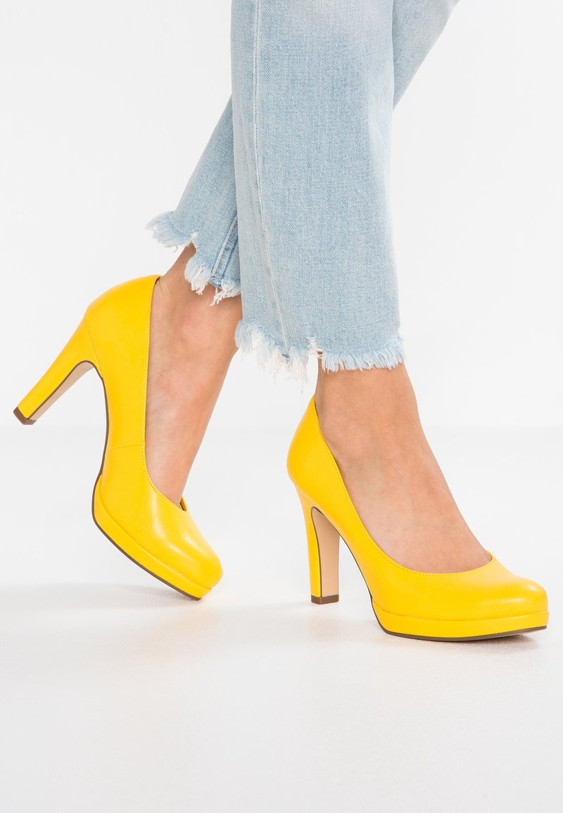 Femme portant un jean bleu clair à ourlet effiloché et des escarpins fermés à talons hauts de couleur jaune vif, debout sur une surface blanche.