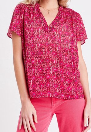 Femme portant un chemisier rose vif à motif floral avec col en V et manches courtes, pantalon rose uni, colliers dorés superposés visibles.