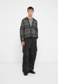Cardigã de malha em padrão xadrez verde com colarinho, combinado com calças cargo pretas largas e sapatos pretos. T-shirt cinza por baixo do cardigã.