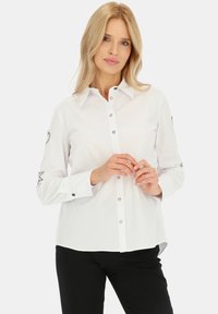 Biała koszula zapinana na guziki z gładkiego materiału z długimi rękawami. Posiada guziki w srebrnym odcieniu oraz ozdobne detale na mankietach. Czarne spodnie.