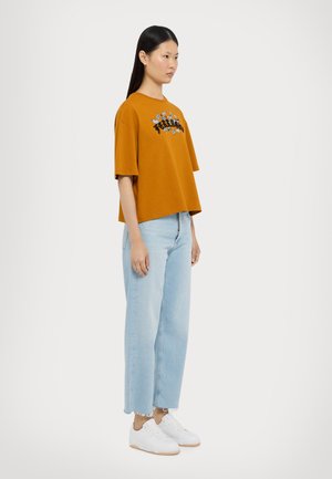 Tricou supradimensional de culoare muștar cu broderie florală, asociat cu blugi drepți de culoare albastră deschisă și teniși albi. Îmbrăcăminte simplă și casual.