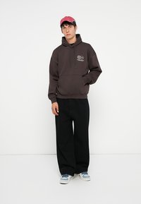 Brun hættebluse med kangaroo-lomme, der har et logo-print. Matchet med løse sorte bukser og hvide sneakers. Inkluderer en lyserød kasket.