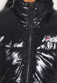 Giacca a vento nera con finitura lucida, colletto alto, chiusura frontale con zip e logo ricamato rosso e bianco sul lato sinistro del petto.