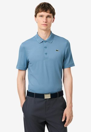 Junger Mann trägt ein hellblaues kurzärmeliges Polohemd mit kleinem Krokodil-Logo und dunkle Hose mit schwarzem Gürtel.
