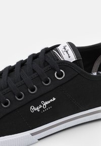 Black canvas sneaker featuring flat black laces, silver eyelets, white rubber sole, and Pepe Jeans logo on the side.