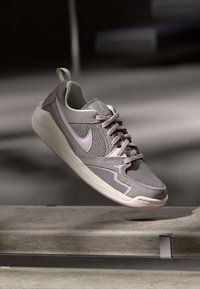Ανοιχτό μωβ και γκρι Nike sneaker με λεπτομέρειες από διχτυωτό υλικό και σουέτ, οι κορδόνες δεμένες, εμφανισμένο στον αέρα πάνω από μια τσιμεντένια επιφάνεια.