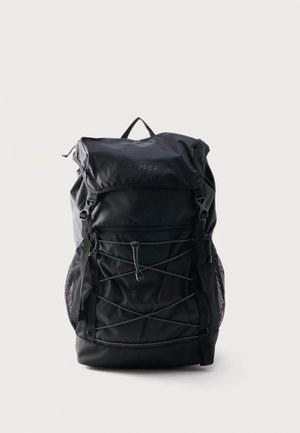 Sac à dos Terrex noir avec rabat supérieur, poche avant zippée, bretelles réglables et détail en corde élastique, posé sur un fond uni.