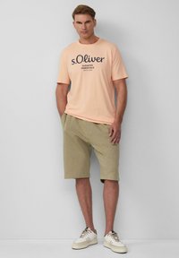 Perzikkleurig t-shirt met korte mouwen en donkere print, gecombineerd met lichtgroene shorts. Model draagt beige sneakers met zwarte accenten. Eenvoudig ontwerp.