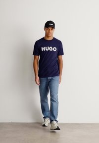 Jūras spēku t-krekls ar lieliem baltajiem "HUGO" burtiem, apvienots ar gaiši ziliem džinsiem un melniem sporta apaviem. Melna cepure ar logotipu noslēdz apģērbu.