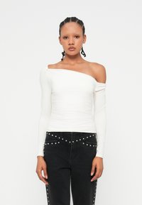 Bela bluza brez naramnic z dolgimi rokavi, prilegajočega kroja in gladko teksturo, v kombinaciji z belimi kavbojkami, ki imajo ujeti okrasni detajli ob šivih.