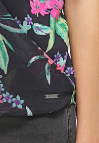 Blusa floral con vibrantes patrones en rosa, verde y azul sobre un fondo negro; presenta un pequeño acento de logo metálico.