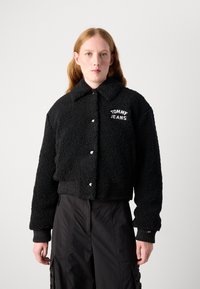 Veste noire courte en tissu pelucheux avec un col, arborant le logo blanc "TOMMY JEANS" et des poignets et un ourlet côtelés. Portée avec un pantalon noir.