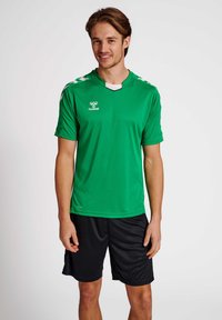 Grünes Sporthemd mit weißen Akzenten und Logo, kombiniert mit schwarzen Shorts. Das Hemd hat kurze Ärmel und ein V-Ausschnitt-Design.
