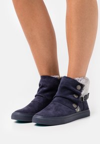 Botas de tornozelo em camurça azul-marinho com forro de pele sintética, apresentando um design com dobra, botões prateados e uma sola de borracha texturizada.
