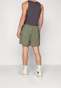 Mies, joka on pukeutunut oliivinvihreisiin shortseihin, tummanharmaaseen ilmanhihaiseen puseroon, valkoisiin sukkiin ja valkoisiin korkeisiin tennareihin, seisoo tasaisella taustalla.