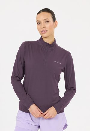 Frau mit zurückgebundenem dunklem Haar trägt ein lila Langarm-Half-Zip-Oberteil und helllila Shorts vor einem weißen Hintergrund.
