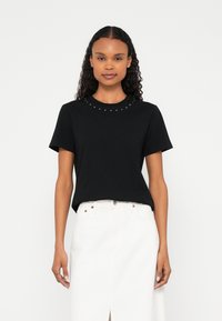 Donna con capelli ricci che indossa una maglietta nera con borchie e scollo tondo e una gonna bianca a vita alta con una spacco frontale, in piedi davanti a uno sfondo bianco.