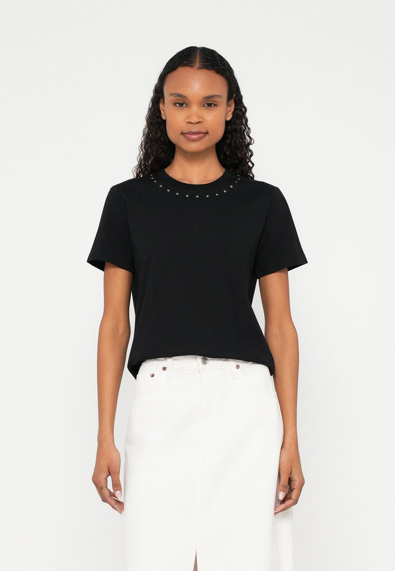 Donna con capelli ricci che indossa una maglietta nera con borchie e scollo tondo e una gonna bianca a vita alta con una spacco frontale, in piedi davanti a uno sfondo bianco.