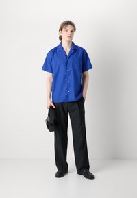 Camisa de manga curta azul com um bolso, combinada com calças pretas. O sujeito segura uma bolsa preta; o conjunto apresenta um ajuste descontraído e um design simples.