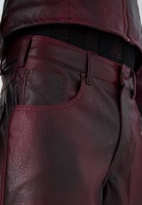 Pantalon en cuir bordeaux avec surface texturée, passants de ceinture et coutures contrastantes. Il présente des poches avant et un design ajusté.