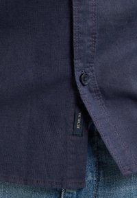 Laivastonsininen pellavapaita, jossa on nappi ja ommeltu helma, ja jossa on sivussa etiketti. Se on kerrostettu denim-kankaan päälle.