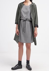 Olivgrön lätt regnjacka över en svartvit mönstrad klänning, åtdragen i midjan med ett bälte. Svarta skor och strumpor kompletterar looken.