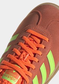 Ruskea mokkanahkainen tennari, jossa on neonvihreät raidat ja oranssit nauhat. Ominaisuuksiin kuuluu teksturoitu kieli ja kumipohja sekä sivussa oleva logotarra.