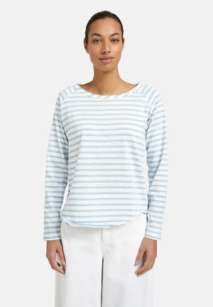 Young woman wearing a light blue and white striped long-sleeve shirt and white pants, standing against a plain light background.