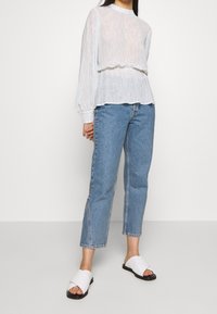 Femme portant un chemisier transparent à motifs blancs à manches longues, un jean bleu coupé avec des fentes sur le côté, et des sandales blanches à enfiler avec des semelles noires.