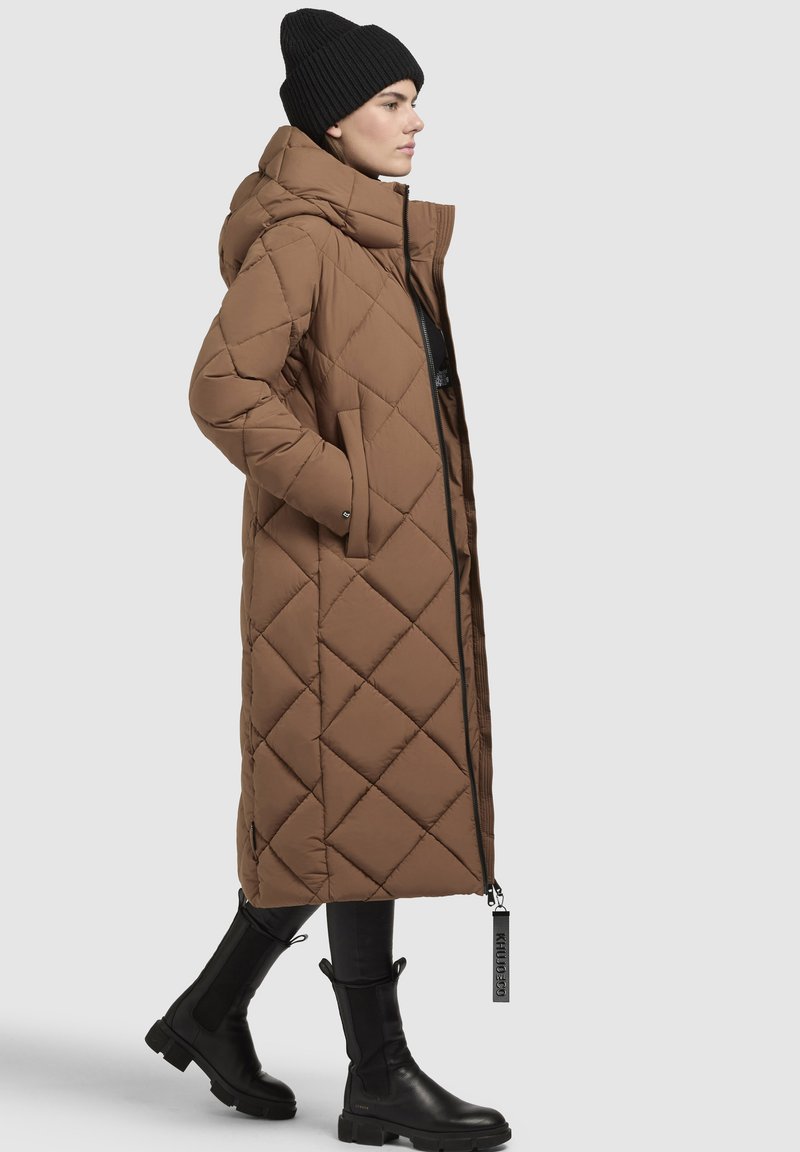 Brauner, gesteppter langer Mantel mit Kapuze, ausgestattet mit einem Reißverschluss, seitlichen Taschen und schwarzen Akzenten, kombiniert mit knielangen schwarzen Stiefeln.